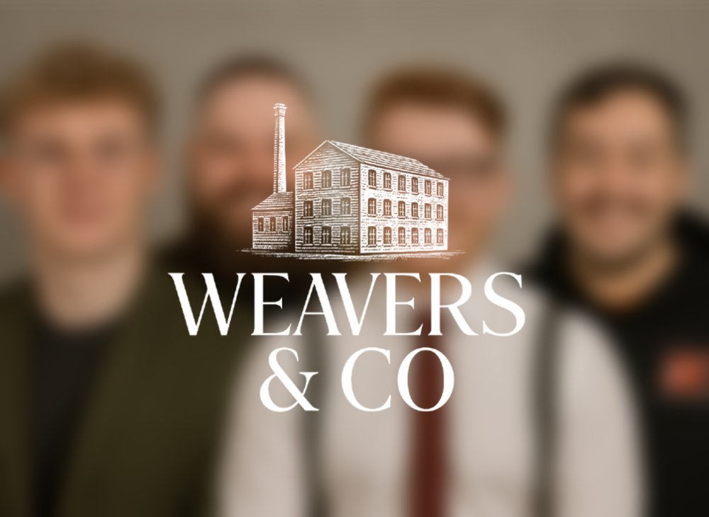 A friendly, professional group portrait of four team members from Weavers & Co, standing shoulder to shoulder in front of a soft gray backdrop. From left to right: a blond man in glasses and an olive blazer, a bearded man in a checkered flannel shirt, a smiling red-haired man in a shirt and tie with suspenders, and a casually dressed man in a black Nike hoodie. The Weavers & Co logo is subtly overlaid at the top, reinforcing the team’s creative and approachable brand identity.